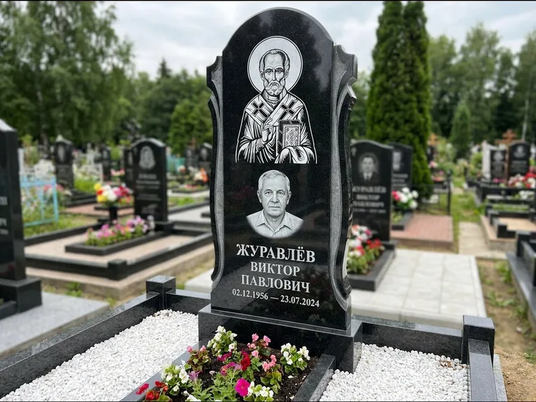 Памятник с гравировкой иконы: стела с образом в полный рост на обороте — гравировка образа Матроны Московской, низкие кусты спиреи в цвету, работа «Лебедева» в Нижнем Новгороде